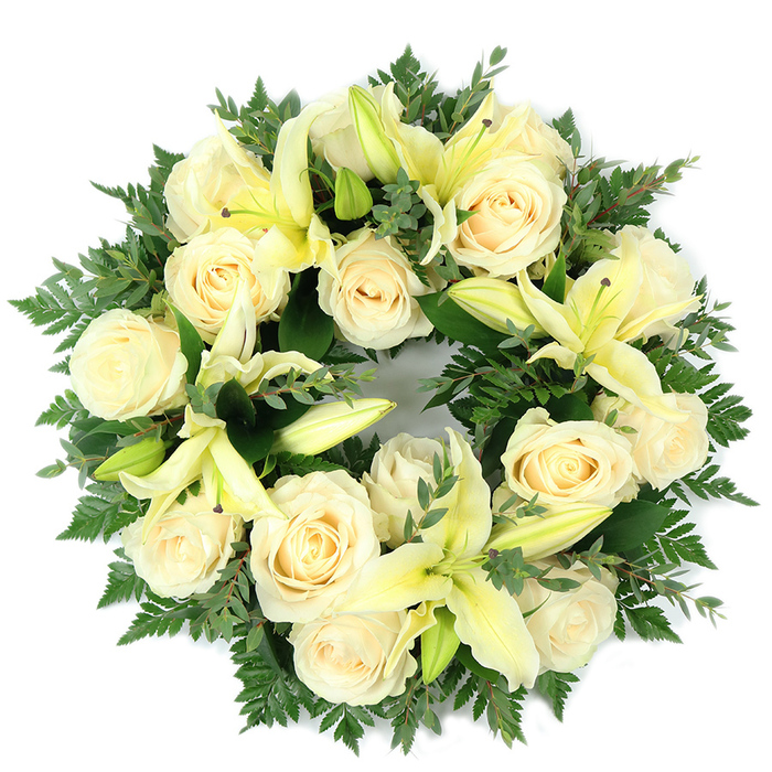 A meticulously crafted circular funeral wreath laid flat and captured against a plain white background, where a constellation of creamy white roses and pale, lemon-tinged lilies forms a serene and full-bodied ring. Approximately fifteen to twenty large roses with compact, spiralling petals provide a luminous, ivory field; interspersed among them, soft yellow trumpet lilies open with graceful curvature while a few remain as poised buds, lending a sense of promise and gentle progression within the composition. The blooms rest upon a generous foundation of varied greenery: delicate, feathery fern fronds soften the wreath's outer silhouette and darker, broader sprigs - reminiscent of eucalyptus - give weight, contrast and a cool green counterpoint to the warm florals. The wreath is arranged with careful symmetry and density so that viewed from above it reads as a continuous, comforting surface, the interplay of textures inviting both visual and tactile imagination. Light is even and mild, bringing out the roses' creamy sheen and the lilies' subtle lemon tones without harsh highlights. There is an understated fragrance implied - the roses' soft powder balanced by the lilies' fresh, slightly citrus edge - which enhances the arrangement's calming presence. Intended as a dignified memorial or sympathy tribute for funeral services, graveside placement, or to offer condolence in the Mill Hill area, this wreath embodies themes of purity, peace and enduring remembrance while reflecting the thoughtful hand of a local florist who understands the need for respectful, composed floral expressions in times of loss.