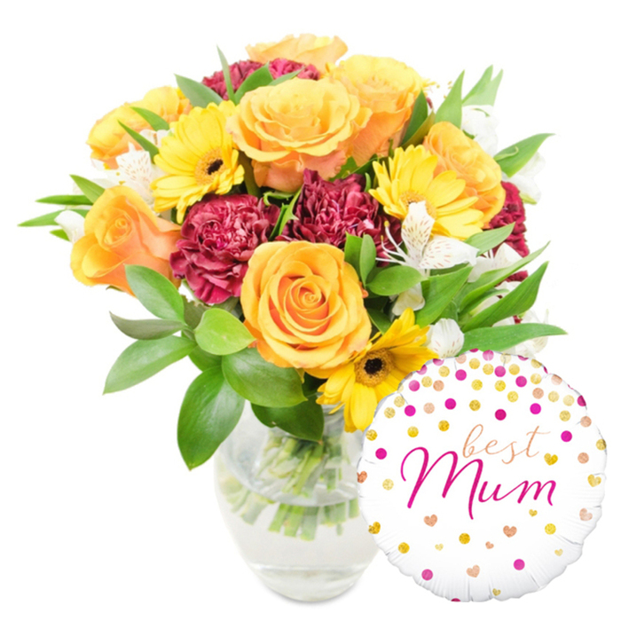 A vivid, celebratory floral composition set against a stark white studio background: the bouquet, arranged in a simple clear glass vase and placed slightly left of centre, is a study in warm contrasts and textures, while a cheerful round foil balloon rests at the lower right, overlapping the vase base. The blooms centre on plump orange roses - their petals swelling outward in soft, concentric curves - surrounded by bright yellow gerbera daisies whose sun-like petals and dark brown centres draw the eye. Deep red to burgundy carnations, tightly ruffled and velvety in appearance, provide a plush, shadowed counterpoint, and crisp white alstroemeria with narrow, freckled petals streak through the grouping like delicate punctuation. A generous accompaniment of glossy green foliage - from broad, reflective leaves to finer, wiry sprigs - both supports the flowers and creates a natural, garden-fresh silhouette; you can see the trimmed stems through the clear water, a quiet indicator of recent arranging. The white foil balloon is sprinkled with gold and fuchsia confetti-dots and tiny hearts; across its centre an elegant fuchsia cursive reads best Mum, with best tucked above the larger, more emphatic Mum. The overall effect is joyful and intimate, perfect for a Mother's Day surprise, a birthday celebration, or a spontaneous thank-you to a beloved mother living near The Ridgeway, Mill Hill Park or Bittacy Hill, where neighbours often choose similar bright, thoughtful combinations to mark special moments.