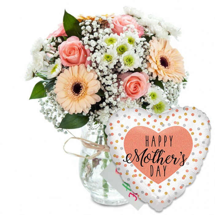 A beautifully styled Mother's Day gift ensemble is displayed against a clean white background, creating a calm, luminous space where every petal, leaf, and reflective highlight can be appreciated. On the left side of the composition, a rounded bouquet of mixed fresh flowers sits proudly in a clear glass jar-style vase. The vase is cinched at the neck with a wrap of rustic twine, a small detail that instantly lends warmth and the feeling of a thoughtful, local Mill Hill touch rather than something mass-produced. The bouquet showcases several softly coloured roses in shades of peach and light pink, their petals unfurling in layered spirals that appear almost satin-like. Intertwined among the roses are two or more sizeable peach gerbera daisies, instantly recognisable by their neat rings of slender petals and pronounced dark brown centres that draw the viewer's gaze inward. Delicate clouds of white baby's breath are scattered throughout the bouquet, adding an ethereal, misty texture, while clusters of small white chrysanthemums, each with a hint of pale green at its core, lend structure and fullness. Sprays of lush green foliage peek between the blossoms, providing contrast and depth, reminiscent of the leafy gardens and tree-lined streets around Mill Hill. To the lower right of the vase, overlapping it slightly, a large heart-shaped foil balloon balances the composition. The balloon's bright white surface is sprinkled with metallic gold and silver dots, resembling festive confetti frozen in place. At its centre, a generous peach-coral heart shape contains the phrase HAPPY Mother's DAY in bold black lettering, combining clean, modern capitals with an elegant script that emphasises the word Mother's. Just behind the flowers and balloon, the corner of a small white greeting card can be seen, reinforcing the sense of a complete, ready-to-give gift. The entire scene evokes warmth, gratitude, and celebration, perfectly suited for illustrating Mother's Day bouquets delivered across Mill Hill, from quiet cul-de-sacs off Bittacy Hill to homes overlooking Mill Hill Park.
