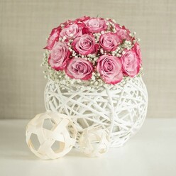 An elegant floral composition is captured as if it were a quiet moment in a Mill Hill townhouse, with a lush bouquet of pink roses and baby's breath displayed in a distinctive white woven vase. At the centre is a generously rounded cluster of soft pink roses, densely packed so that only their heads are visible, creating a cushion of petals. Each rose reveals a subtle ombré of colour, the heart of the flower starting in pale blush and shifting toward a deeper, near-fuchsia pink along the outer edges, giving the bouquet depth and movement. Between the roses, fine stems of white baby's breath weave through like mist, their clusters of tiny blossoms lending an ethereal, cloud-like texture that evokes wedding aisles and first dates along the quiet streets off Bunns Lane. These blooms sit in a spherical container crafted from white-painted, interwoven strands that resemble wicker or rattan, forming an open lattice that allows light to pass through. The vase feels both contemporary and handmade, as though sourced from a local craft market and paired thoughtfully with fresh flowers by an experienced Mill Hill florist. Resting on the sleek white surface in front of the vase are two decorative balls made from light, natural fibres, their strips crossing in loose, geometric patterns. One sphere is noticeably larger, and together they echo the vase's texture while adding a minimalist design detail. The backdrop is a softly textured wall in a warm beige tone, catching gentle, even light that enhances the softness of the pinks and whites without creating strong shadows. The arrangement exudes calm sophistication, ideal for a bedside table, a hallway console, or a small celebration at home when you want to express affection in a quietly luxurious way.