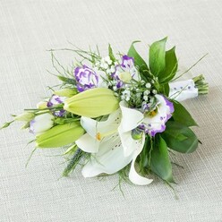 A refined lily and lisianthus bouquet rests at a diagonal on a pale beige, woven-textured cloth, the kind of serene setting you might imagine on a dressing table before a Mill Hill wedding. The composition is dominated by several open white Oriental lilies, their broad, silky petals arching outward in perfect symmetry. Each lily reveals a cluster of yellow-orange stamens dusted with pollen, surrounded by faint reddish freckles that lead the eye gently into the throat of the flower. Alongside them, fresh chartreuse-green buds stand poised to open, their smooth outer surfaces contrasting with the softness of the full blooms. Woven among the lilies are striking Eustoma flowers, also known as lisianthus, with their delicately frilled, cup-like petals. Their coloration shifts from clean white centers to vivid purple edges, with subtle washes of lavender and violet appearing where the tones blend. Fine sprays of white baby's breath drift between these focal blooms, creating a sheer, cloud-like texture that gives the bouquet an airy, romantic lightness. Surrounding everything is a frame of greenery: broad, glossy leaves that provide structure; softer, feathery fronds; and a few more angular green accents that add a modern touch. At the base, the stems are carefully bound in a smooth white satin ribbon, detailed with tiny pearl-style beads that form a gentle line along the exposed end, reinforcing its ceremonial feel. The soft, even lighting in the image highlights the freshness of the petals and foliage without casting deep shadows, allowing each layer of texture and colour to be clearly sensed. This bouquet feels ideal for a bride walking from Mill Hill East station to a nearby ceremony, as a thoughtful anniversary arrangement, or as an elegant centrepiece delivered locally from Flowers Mill Hill to a family gathering near Arrandene Open Space.