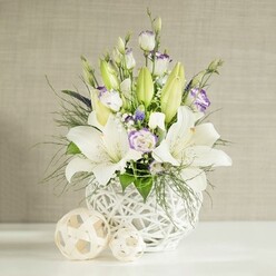 A sophisticated white and lavender-inspired floral design is displayed as though set on a wedding reception table in Mill Hill, bathed in soft, diffused light. The flowers are gathered into a rounded white vase with an intricate open lattice, reminiscent of woven wicker but shaped with modern elegance. From this airy container, tall stems of white lilies rise gracefully, some in full bloom with wide, trumpet-like petals curving outward to reveal pale green throats and lightly dusted stamens, others held as elongated green buds reaching for the light.   

Around the lilies, lisianthus flowers introduce a romantic softness. Their petals are predominantly white, with shy hints of lavender-purple along the edges or in the centres, suggesting a hand-painted touch. Some lisianthus are fully open, forming layered rosettes, while others remain in slender, droplet-like buds that add vertical interest. Tiny white filler blossoms, likely Gypsophila, are sprinkled throughout, and a few deeper purple florets nestled between the stems give the design gentle visual depth. A variety of green foliage, from fine, feathery sprigs to broader leaves, fills out the silhouette, some stems arching over the rim of the vase for a natural, garden-inspired feel.   

On the white tabletop before the vase sit two light beige woven spheres, their open-work structure reflecting the vase's pattern and tying the composition together. Behind the arrangement, a soft, light beige wall with subtle horizontal texture recedes quietly, allowing the flowers to take full focus. The overall mood is serene and polished, perfect for wedding breakfasts at Mill Hill Golf Club, intimate anniversary dinners at home, or thoughtful get-well arrangements delivered locally by Flowers Mill Hill.