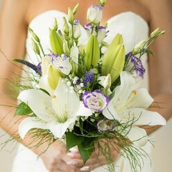 In a light-filled studio in Mill Hill, a bridal bouquet takes form in the hands of a skilled florist-a vision of romance, tailored for a wedding steeped in North London charm. Towers of creamy white Oriental lilies unfurl at the core of the arrangement, each bloom luminous, flecked gently with maroon, and crowned with soft yellow stamens that signify devotion. White lisianthus blooms, their petals like silk edged in faint violet, are nestled closely to suggest both tenderness and whimsy, some bursting open while others remain in bud, packed with hope for the future. The deep purple spikes of Veronica snake upward, bestowing the design with rich intensity, complemented by light clouds of baby's breath that give the whole bouquet an ethereal softness. A foundation of deep green lily leaves and wispy asparagus fern provides dynamic movement, mirroring the layered hedgerows that line Mill Hill's quiet lanes. The bride's manicured hands, finished in pale pink polish, delicately hold the stems, and a strapless white wedding dress serves as a serene, blurred backdrop. Every element has been chosen with care and an understanding of Mill Hill's discerning clientele, resulting in an arrangement that radiates freshness, subtle luxury, and the promise of lasting happiness-perfect for same-day delivery throughout the NW7 area and neighbouring London villages.