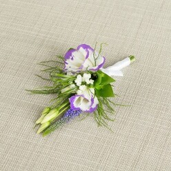 Set against a soft, woven light beige fabric that looks like fine linen, this floral boutonniere feels as if it has been paused in a moment of calm just before a special occasion in Mill Hill begins. Two white lisianthus flowers form the focal point, each petal gently ruffled and brushed at the edges with a vivid, velvety purple, creating a painterly halo that frames their open centres. Around them, tiny clusters of white filler flowers hover like a delicate mist, bridging the space between the main blooms and the surrounding greenery. Rising vertically through the composition is a narrow stem of muscari, or grape hyacinth, its small, tightly packed bell-shaped florets a deep, cool purple that adds texture and depth, reminiscent of the jewel-like blossoms seen in spring borders along the paths near Mill Hill East. Two pale green buds sit quietly at the edge, still closed, embodying a sense of anticipation that mirrors the emotions of a groom or prom guest preparing for the day. Fine, feathery greenery, similar to asparagus fern, arcs gently around the flowers, softening their outline and lending a whisper of meadow-like movement. At the base, the slender stems are meticulously bound and smoothed under a wrap of crisp white satin ribbon, finished with a tiny, tidy bow that announces its purpose as a lapel accessory. The overall design is classic yet gently romantic, perfectly suited to a wedding at a Mill Hill church, a celebration in a local restaurant, or any formal event where a subtle touch of floral elegance and local character is desired.