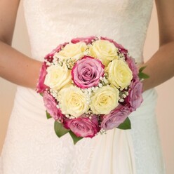In this intimate portrait, captured in Mill Hill's gentle daylight, a bride stands serenely with her bouquet held at the center, forming a symphony of roses that exudes both refinement and emotion. The arrangement is masterful: creamy lemon-yellow roses gathered tightly with bursts of fuchsia pink, their petals blushing ever-so-slightly with hints of mauve and lavender-a color harmony evocative of summertime blooms found in Mill Hill's community gardens. Fine stems of baby's breath interlace with the roses, casting a delicate, cloud-like texture across the bouquet and lending visual depth. Lively green foliage peeks discreetly from beneath, referencing the abundant greenery that lines local streets near Mill Hill Broadway. The bride's bare arms securely frame the arrangement, while her gown, soft ivory in tone, boasts intricate lace patterns that shimmer in the even light, the skirt gently pooling in quiet, graceful folds. The lighting reveals the vibrancy and freshness of each flower and the craftsmanship of the lace, ensuring every fine detail is appreciated. Behind, a subtle, blurred background-light and understated-keeps the focus on the bouquet and the romance of the moment. Perfect for those planning a celebration in Mill Hill or nearby green spaces, this bouquet is the living centerpiece of a cherished tradition. Created by experienced florists with an understanding of local style, it captures the essence of bridal sophistication, the promise of new beginnings, and the elegance of a special day in this beloved North London community.