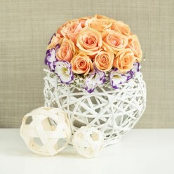 On a sleek white tabletop, a beautifully balanced floral centerpiece glows with quiet elegance, as if styled for a wedding reception in one of Mill Hill's hidden venues or a family celebration in a light-filled dining room near Mill Hill Park. The focus is a lush, rounded mound of roses in shades of peach, apricot, and soft salmon, the flowers packed closely so their silky petals form a smooth dome that feels almost cushion-like. Each rose is fully opened, revealing generous spirals that draw the eye inward and suggest warmth and affection. Circling the base of this rose dome is a graceful band of lisianthus blooms, their white petals frilled at the edges and finely tipped in a rich purple hue that deepens toward the rim, like ink on handmade paper. Threaded among them are delicate sprigs of white gypsophila, tiny blossoms creating a misty, lightweight texture that softens and connects each element. The bouquet is anchored in a white woven container, broad and gently bulbous, made from intersecting strands of rattan or wicker that form an open, lattice pattern. Light slips through the gaps, casting subtle shadows that emphasise its hand-crafted character, much like artisan pieces discovered in small Mill Hill boutiques. In the foreground, two decorative spheres rest on the tabletop-one larger, one smaller-each fashioned from pale wooden strips looped and intertwined into open, airy orbs that echo the vase's texture and add sculptural interest. Behind the arrangement, a softly textured beige backdrop, reminiscent of natural linen, brings warmth and a hint of rustic charm, allowing the flowers to stand out clearly. The entire scene conveys gentle romance and refined calm, perfectly suited to wedding tablescapes, engagement parties, or elegant home décor across Mill Hill and the surrounding area.