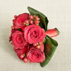 A striking, compact bouquet of deep red roses and berries lies gracefully on a pale beige, textured fabric, like a corsage or bridesmaid posy set down for a moment between photographs at a Mill Hill wedding. At the heart of the image, several lush roses in full bloom form a tight, rounded cluster, their velvety crimson petals spiralling inward to darker, shadowed centers. The petals appear soft and cool to the touch, with subtle variations of scarlet and wine red that suggest depth and movement, as if each bloom is still quietly opening. Threaded among these roses are small clusters of rounded Hypericum berries in rich shades of red and rosy pink, their smooth, glossy surfaces catching the light and adding an element of playful texture. Surrounding and supporting the flowers, broad, dark green leaves are artfully folded and curved, their clean lines and polished finish giving structure to the arrangement, rather like a tailored collar on a well-fitted jacket. At the base, mostly hidden but gently visible at one side, a richly coloured red satin ribbon binds the stems, its soft sheen mirroring the luxury of the petals and suggesting the careful hands that tied it. The background fabric, in a warm cream-beige tone with delicate texturing, keeps the composition calm and understated, allowing the intense reds and greens to stand out. The overall mood is romantic and polished, ideal for imagining same-day flower delivery around Mill Hill Broadway, a bridesmaid bouquet carried through local churches, or a refined floral accent for an intimate dinner at home, crafted with quiet precision and emotional warmth.