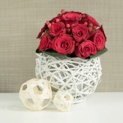 A richly textured wedding-style centerpiece fills the frame, composed of a dense, rounded bouquet of red roses that looks almost like a perfectly sculpted floral dome. The roses themselves are in various stages of opening, from tight buds that hint at their inner petals to fully unfurled blossoms whose velvety layers spiral inward, catching the soft light in subtle highlights and shadows. Threaded among the roses are clusters of deep red Hypericum berries, each tiny orb smooth and glossy, offering a pleasing contrast to the ruffled rose petals and adding a sense of rhythm and movement to the design. A discreet collar of dark green leaves cradles the base of the bouquet, the foliage only just visible but helping to define the shape and lend a natural depth to the arrangement. All of this beauty is held in a distinctive spherical vase made from white-painted wicker or twigs. The vase is crafted as an open lattice, with the intertwined branches forming a airy, sculptural sphere that allows glimpses of the interior and plays gently with light and shadow. It sits on a pristine, white tabletop that reflects some of the arrangement's colour and structure, reinforcing the clean, modern aesthetic. To the left, two smaller decorative accents-wicker balls made from pale, natural wood-rest on the same white surface. The larger one lies slightly in front, with the smaller ball behind, both echoing the woven texture of the vase and adding a soft, organic note. The background is a softly blurred, neutral-toned wall, similar to the calm, contemporary interiors found in Mill Hill homes overlooking local green spaces like Mill Hill Park. The overall mood is warm and romantic, suggesting a carefully curated centerpiece ready for a wedding reception table or a celebratory meal in this peaceful corner of North London.