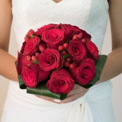 This luxurious bridal bouquet, photographed just outside Mill Hill Broadway, becomes an ode to classical elegance and heartfelt celebration. The compact, full-bodied composition features sophisticated deep red roses-each velvety petal unfurling in a choreographed swirl of romantic intensity, their heady fragrance invoking promises exchanged in quiet corners of Mill Hill's lush gardens. Sprinkled within the red blooms, bright, smooth hypericum berries create a lively, tactile accent, lending playful visual punctuation to the bouquet's poetic arrangement. A collar of broad, glossy green leaves embraces the bouquet, reflecting the natural beauty of nearby Arrandene and Bittacy Hill parks. Tender hands support the arrangement, showcasing both the florists' attention to detail and the bride's anticipation. Rich lace and gentle draping of the wedding gown add an aura of tradition. The lighting brings forward every hue and subtle highlight, while the softly muted background enhances the bold reds and fresh greens. An ideal choice for brides seeking a blend of bold passion and enduring tradition, this bouquet from Flowers Mill Hill echoes the vitality of local celebrations and the skill of North London floral design.