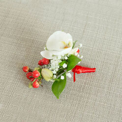 Laid carefully on a pale, woven linen backdrop in a soft beige tone, this elegant boutonniere is captured from above as though it has just been placed on a dressing table before a ceremony in Mill Hill. At its centre sits a flawless white Phalaenopsis orchid, the petals wide and velvety, curling outwards with a gentle movement that feels almost like a breath. In the middle of the bloom, a touch of buttery cream and muted orange glows in the throat, giving the orchid a warm, luminous heart. Around it, sprays of delicate white baby's breath form a fine halo, their tiny star-shaped blossoms creating a cloud-like softness and an almost whisper-light texture. To one side, a cluster of round, glossy red berries gleams against the neutral background, adding a festive, jewel-like accent that brings to mind winter weddings and Christmas services near Mill Hill Broadway. A few paler green berries are tucked alongside, hinting at newness and natural variation. The arrangement is framed by rich green leaves, some broad and pointed, others slender and more elongated, each leaf surface catching subtle highlights from the ambient light. At the base, the stems are bound tightly in a rich red satin ribbon, wrapped smoothly and tied with a small, neat knot, its short tails lying gracefully across the fabric. The design feels carefully composed yet effortless, as if prepared by a local florist who understands the quiet significance of a groom's buttonhole. Perfect for weddings, proms, or black-tie events across Mill Hill, this small piece of floral artistry offers a calm, confident touch of nature that accompanies the wearer through their most meaningful moments.