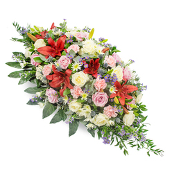 Against a clean white background, this long, lush casket spray appears as a river of flowers and greenery, carefully composed by an experienced Mill Hill florist. The piece is elongated and gently tapered, designed to sit along the length of a coffin and be appreciated from every angle during a farewell service. Deep red lilies, with their large, star-shaped petals and slightly recurved tips, form powerful focal points through the centre and edges, their rich hue suggesting strength, devotion, and heartfelt emotion. Nestled between them are creamy ivory and soft pink roses, some tightly furled like promises, others fully opened to reveal their layered hearts, expressing love and cherished memories. Pale pink carnations, their petals finely ruffled, lend softness and volume, filling the arrangement without feeling heavy. White chrysanthemum-style blooms and small daisy-like flowers with yellow centres lighten the palette, echoing a sense of hope and peace. Threaded throughout are cool blue-purple thistles and tiny clusters of lavender-blue filler flowers, adding texture and contrast, evoking wildflowers you might encounter on a quiet walk near Mill Hill's fields and green lanes. The flowers are cradled in a rich framework of greenery: broad, glossy leaves for structure, softer fern-like sprigs for a feathery finish, and trailing foliage that gently extends beyond the main body of the spray. The combination of warm reds and pinks with cooler purples and greens creates a balanced, contemplative mood, ideal for expressing sympathy and respect at a funeral or memorial gathering in and around Mill Hill.