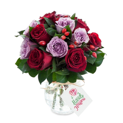 This lush, round bouquet pairs intense red roses with pale lavender companions in a composition that feels both dramatic and tender. Deep crimson and burgundy roses form a luxurious core, their layered petals folding inward like velvet, while soft mauve and lilac roses breathe a cool, calming note through the arrangement. Speckled throughout are small, glossy clusters of red-orange rose hips that add a lively punctuation and tactile contrast to the soft petals-their subtle firmness invites a gentle touch. Dark green leaves, perhaps Ruscus, weave beneath and between the blooms, creating a sculpted backdrop that accentuates every shade. The flowers are presented in a bulbous glass vase with a crackled, mercury-glass finish that picks up highlights and creates an antique, reflective halo around the stems. A length of natural twine is tied simply at the neck, from which a white rectangular tag dangles, waiting for a short, personal message. Photographed against a pristine white background, the bouquet's colours appear richer, the textures more immediate, and the whole arrangement exudes a sense of considered elegance. Imagine receiving this on a quiet morning in Mill Hill, perhaps after a walk by Dollis Brook, its scent faintly floral and green, the arrangement offering reassurance, admiration, or a romantic promise. It reads as a premium, thoughtfully crafted gift suitable for anniversaries, birthdays, or moments when you want to show someone they are seen and cherished.