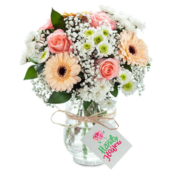 Imagine sunlight filtering through the tall trees bordering Mill Hill Broadway, illuminating a bouquet whose soft pastels and lush forms evoke peace and celebration. Blooming at the centre, peach-pink roses radiate a gentle sweetness, their spiral petals opening beside the lively faces of peach gerbera daisies highlighted by rich brown centers. Surrounding them, white chrysanthemums with vibrant green eyes add clarity and understated cheer, while delicate sprays of baby's breath drift between the larger flowers like soft clouds. Vibrant eucalyptus leaves add texture and a hint of woodland freshness, a reminder of the gardens and walks that define Mill Hill's green spaces. The bouquet is gathered in a crystal-clear, elegantly textured glass vase, its water and stems visible as a testament to expert craft and freshness. Wrapped at the neck with rustic twine, a tidy bow supports a rectangular white tag-blank, ready for your own special message. The overall effect is composed yet relaxed, suitable for birthdays or as a gesture of thanks along Page Street or at the heart of Mill Hill. It's an arrangement that feels equally at home in a sun-filled kitchen or gracing the tables of a family gathering, echoing the serenity and beauty of its local roots.