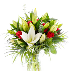 A study in contrast and quiet movement, this bouquet centres on a single expansive white Oriental lily whose pristine petals unfurl to reveal pale yellow stamens and a tender green pistil-the heart of the composition. Encircling that focal bloom are numerous lily buds in soft lime and buttery yellow, their promise of more flowers lending buoyancy and volume. Between the lilies, clusters of small red roses and rosebuds punctuate the palette with velvet depth; they sit like secret letters tucked into a garden, their crimson still tightly curled but eager to open. The arrangement's foliage is layered and textural: glossy, broad leaves in deep forest green form a strong backdrop; longer, palm-like fronds sweep gracefully outward in emerald tones, giving the bouquet a sense of wind-swept motion; and rounded, silvery eucalyptus leaves thread through the stems, offering a muted, cooling counterpoint. A plain cylindrical glass vase holds the stems in clear water, letting the pale green shafts show through and reinforcing the design's fresh, understated clarity. Bathed in bright, neutral studio light against a white background, the colours and surfaces-the lily's satiny petals, the rose's velveteen curves, the eucalyptus' matte sheen-read with elegant precision. The scent, if you could imagine it, would be a gentle blend of sweet spice and heady rose. As a Mill Hill florist familiar with local tastes-whether for gatherings near Mill Hill Park or intimate moments in Mill Hill Village-this composition feels suited to celebrations, anniversaries, or a polished sympathy tribute.