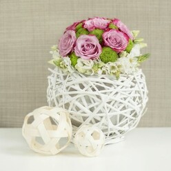 A serene, studio-style image showcases a carefully crafted floral centerpiece, the kind that might sit at the heart of a wedding breakfast table in Mill Hill's leafy suburbs. The flowers form a generous, domed bouquet, composed primarily of full-bloom roses in soft pink and muted lavender tones. Their petals spiral outward in plush layers, with many blooms displaying slightly deeper pink at the edges, creating gentle visual interest. Nestled amid these romantic roses are bright green button chrysanthemums, their compact, round heads and fine, radiating petals offering both vibrant color and a tactile, almost playful texture. Around the lower edge of the dome, tiny white filler flowers cluster and peek out, lightening the overall silhouette and adding a delicate, cloud-like softness. The flowers rise from a distinctive white vase made of intertwined rattan-style strands, woven into a hollow spherical form with open gaps that let the background softly filter through. This airy structure has a contemporary yet organic character, evoking the calm, modern homes around Mill Hill Broadway. To the left of the vase, two decorative spheres in natural pale wicker rest on the same smooth white tabletop; one is noticeably larger, and both feature an open, crisscrossing lattice pattern that echoes the vase's weaving and brings balance to the arrangement. The backdrop is a warm, neutral beige with a subtle fabric texture, keeping the focus firmly on the floral artistry. Soft, even lighting caresses every petal and strand, conveying a mood of understated sophistication and romance, ideal for weddings, anniversaries, or intimate celebrations across Mill Hill.