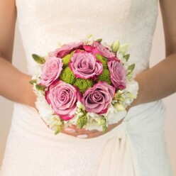 Evoking a sense of refined joy, this striking bridal bouquet is the centrepiece of a Mill Hill wedding morning-lush and thoughtfully arranged by local hands. An ivory lace dress serves as the backdrop, its pronounced floral motif and graceful drapery conjuring images of gatherings at Mill Hill's historic halls and leafy parks. Dominating the bouquet, velvety roses in dusty mauve and soft pink bloom with romantic vibrance; their fuchsia-edged petals catch glimpses of morning sunlight, suggesting both exuberance and intimate calm. A cluster of vibrant lime-green pom chrysanthemums nestles between the roses, their round, gently dew-kissed texture adding an uplifting freshness that celebrates nature's renewal in north London. Around the outer edge, a crown of cloud-white flowers-possibly freesias or clusters of tiny spray roses-prescribes elegance and timeless beauty, their presence softened by sprigs of slender greenery. The bride's meticulously finished nails, touched with rosy pink polish and diamond shimmer, cradle the arrangement with care. Every stem and colour choice is expertly orchestrated for couples looking to infuse their special day with meaning, artistry, and the leafy spirit of Mill Hill, as though this bouquet were gathered fresh from the paths winding past St Joseph's College or the green reaches of Bittacy Hill.
