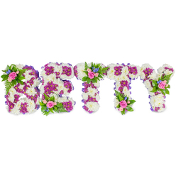 An elegant sympathy tribute meticulously crafted to spell BETTY in uppercase letters, photographed on a pristine white background so every floral detail is visible. The body of each letter is built from dense, cream-white chrysanthemum heads that create a plush, reverent surface - tactile and soft to the eye. Within those white fields, a secondary pattern of deep magenta-purple chrysanthemums forms inner contours and visual accents, adding depth and a luminous contrast that gently defines each letterform. Along the outer flanks a narrow purple ribbon or fabric edging adds a subtle seam, lending the composition a finished, funeral-appropriate refinement. Small bouquet accents punctuate the design: groups of pale pink roses, some newly opened and others still in bud, are set at focal points such as the top of the B, the centre crossbar of a T, and the upper arm of the Y. These roses are accompanied by little sprays of bright blue blooms - possibly delphiniums or small cornflowers - and fresh green fern fronds that provide both colour harmony and a soft, natural frame. The overall mood is dignified and comforting; the cool whites quiet the scene, the purple conveys respectful remembrance, and the pink roses bring a personal tenderness. As an experienced Mill Hill florist, I picture this arranged for a chapel service or a celebration of life near Mill Hill Park, a piece intended to speak for those moments when words fall short and a thoughtful, handcrafted floral name can convey love, tribute and farewell.