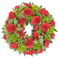 Set against a clean, white backdrop, a circular memorial wreath commands attention with its rich contrast of crimson red and lime green. The ring is completely covered in flowers and foliage, creating a sense of fullness and care that would feel at home in a thoughtful farewell service in Mill Hill. Red carnations dominate the design, arranged closely together so that their layered, ruffled petals seem to overlap like gently folded fabric. Their colour is a deep, dignified red that suggests affection, honour, and gratitude. Nestled consistently between them are lime green button chrysanthemums, each bloom perfectly rounded, like little green cushions. The tightly packed petals of these mums give them a smooth, almost velvety look, bringing lightness and a modern touch to the traditional carnations. The blossoms are supported by an abundant collar of greenery: dark emerald fern fronds fan outward, adding depth and structure, while variegated leaves with pale, creamy edges dance between them, breaking up the darker tones. Where the foliage gathers more thickly, a striped ribbon in soft and deeper greens winds through in gentle loops, echoing the nearby leaves so that it feels part of the natural flow rather than a separate accessory. The circular shape is symmetrical and balanced, every section echoing the next, like a visual reminder of life's ongoing cycle. This wreath could be imagined resting at a graveside near Mill Hill Cemetery, standing beside a photograph in a service at a local church, or placed in a reception room overlooking the green spaces around Mill Hill, conveying gentle comfort and presence during a time of loss.