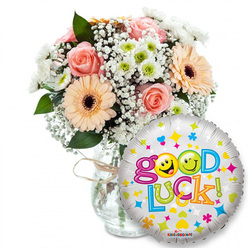 This image shows a thoughtfully composed 'Good Luck' gift set arranged on a crisp white background, the kind of uplifting package we prepare for neighbours across Mill Hill and surrounding streets. On the left a full bouquet nestles in a clear glass vase tied neatly with rustic twine; the blooms are a soft, harmonious mix of light pink roses and peach-toned Gerbera daisies whose darker centres give them weight and character. Fine sprays of white baby's breath (Gypsophila) thread gently through the composition like lace, while compact white-and-lime chrysanthemum clusters introduce a bright, modern accent. Dark green foliage provides contrast and a living structural frame so the pastel shades read clearly against the vase's glass.

Taking the foreground on the right, a round metallic foil balloon gleams with a silver-white sheen and a scattering of colourful celebratory motifs-stars, hearts and confetti-like dots in blue, pink, yellow and green. The bold phrase Good Luck! curves across the balloon in playful, multi-coloured letters; the two 'o's in Good are replaced by two sunny yellow smiley faces-one winking, the other pulling a playful face-bringing an immediate smile. The image feels like a personal send-off: under soft even lighting it radiates warmth and support, ideal for a student heading to exams, a friend starting a new job, or anyone in need of encouragement. I picture this being delivered from a local Mill Hill florist to homes around The Broadway or Mill Hill Park with care and quiet pride.