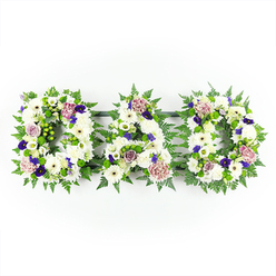A beautifully composed sympathy arrangement spells out DAD in three distinct floral letters, each one constructed so thoroughly from flowers that the underlying frame disappears beneath a cloud of petals. The letters are laid out side by side in a straight horizontal row, set against a simple white background that lets the flowers speak without distraction. The main body of each D and the central A is packed with fresh white chrysanthemums and daisy-like blooms, their rounded, multi-layered petals overlapping to create a soft, velvety texture reminiscent of a comforting embrace. Subtle variations in the centres of the white flowers, some slightly creamy, some with gentle green eyes, lend natural interest while keeping the palette serene. Embracing the letters is a collar of lush greenery-fine strands of fern and glossy green leaves that trace their contours and ensure the word reads clearly. Threaded among the whites are islands of colour: deep violet and rich purple blossoms that could be pansies, lisianthus, or spray carnations, balanced with muted lavender-mauve roses and frilled carnations whose petals look just beginning to unfurl. The result is a gentle play of cool and warm tones, suggesting both sorrow and enduring affection. The lighting is steady and bright, like soft daylight streaming through chapel windows near Mill Hill Broadway, illuminating every nuance of the blooms. This is the sort of personalised floral lettering families in Mill Hill might choose when arranging a funeral or memorial, perhaps to rest atop the coffin during a service or to place later as a lasting tribute. It communicates the word DAD not only in letters, but in the language of flowers, with grace, respect, and quiet love.