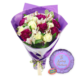 Photographed against a crisp white backdrop, this graduation bouquet and balloon pairing feels like a snapshot of celebration ready to be delivered across Mill Hill. The bouquet stands tall, wrapped in a deep purple paper that hugs the stems snugly before flaring out in neat folds around the flowers, giving the design a polished, ceremonial look. Around the base, a decorative green and white ribbon-possibly patterned with subtle detail-ties the wrapping, evoking the sense of a sash or honour cord worn at a graduation ceremony. Rising from this base is a vibrant mix of blooms. Creamy white roses, their petals tightly coiled into classic spirals, bring an air of elegance and calm. Beside them, clusters of deep fuchsia or magenta blooms, likely carnations or chrysanthemums, burst forward with ruffled, multi-layered petals, adding energy and texture that mirror the excitement of results day near Mill Hill Broadway station. Small white filler flowers, similar to baby's breath, are scattered throughout, their tiny blossoms creating a gentle cloud between the larger blooms. Lush green foliage, in varied shapes and tones, spills out around the flowers, providing a lively natural frame that calls to mind the green spaces and gardens around Mill Hill Park. To the lower right of the bouquet, a round purple Mylar balloon rests lightly on the white surface. Its metallic sheen catches the light, and across the centre, in flowing white script, are the words "To a Loving Graduate," transforming the image into a clear tribute to academic success and family pride. A small oval tag or sticker with green and white details sits beside the balloon, hinting at professional presentation and careful curation. The overall mood is uplifting and hopeful, capturing the moment a proud relative might choose a congratulatory gift for a new graduate in Mill Hill.