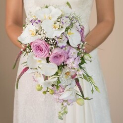 Bathed in soft light reminiscent of early mornings along Mill Hill's The Ridgeway, a bride's arms encircle a lush, cascading wedding bouquet-a testament to the community's love for classic romance and refined floral design. Her gown, detailed with fine lacework and elegant ivory hues, frames an arrangement where creamy white Phalaenopsis orchids take centre stage, their broad petals gleaming serenely, golden throats catching the warm glow. Around them, blush pink and lavender roses-plush, fragrant, each petal luxuriant-create a symphony of colour, texture, and movement. Wisps of baby's breath spiral through the florals, offering a frothy, airy counterpoint echoed by sprigs of lavender stock reminiscent of local hedgerows. Magenta strands of feathery amaranthus cascade downward, their unique form lending drama and a touch of wild grace, as if inspired by Mill Hill's green open spaces. Glossy, fresh foliage and tight buds peek from between the blooms, suggesting new beginnings and community vitality. The subtle background draws focus to the bouquet's artistry, its colour story resonating with the quiet sophistication and hopefulness of Mill Hill ceremonies. Crafted with care and expertise by Flowers Mill Hill, this arrangement becomes more than flowers-it's a symbol of the joy and anticipation cherished by NW7 couples taking their first steps into married life.