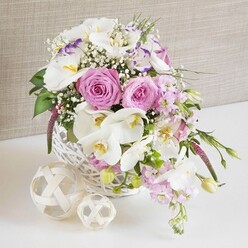 An opulent floral centerpiece overflows from a white wicker basket, captured in gentle, flattering light that feels like a late afternoon reception in Mill Hill. At the core of the design, several large pink roses are fully open, their petals forming intricate spirals that create a sense of depth and romantic allure. These classic blooms are paired with sweeping white Phalaenopsis orchids, whose broad, smooth petals and warm yellow centers lend a sophisticated, modern bridal feel. Nestled among them, white Lisianthus flowers, some brushed lightly with lavender along their frilled edges, offer a delicate, almost porcelain-like texture. Slender spires of pale pink stock emerge and then softly cascade from the basket, giving the impression of blooms gently spilling across a wedding table or family celebration near Mill Hill Broadway. Fine sprays of baby's breath weave through the arrangement, filling any gaps with tiny, cloudlike white blossoms that lighten the overall composition. Pockets of fresh greenery appear between the flowers, with leaves that curve and extend just beyond the rim of the basket, echoing the relaxed beauty of nearby green spaces such as Mill Hill Park. The white wicker basket, gently rounded and finely woven, sits on a clean white surface that reflects subtle hints of pink and green from above. To one side, two decorative spheres created from intertwined natural wood fibres rest in casual harmony, their pale caramel tones balancing the crisp white of the basket. A soft beige, textured fabric background lies quietly behind, offering a neutral canvas that allows every petal and leaf to stand out. The arrangement feels both polished and inviting, perfectly suited to wedding tables, engagement dinners, or refined home entertaining in the Mill Hill area.