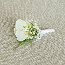 A refined white orchid boutonniere is laid carefully on a neutral, light tan fabric that has the natural, woven look of linen or burlap, evoking the relaxed yet polished style often seen at Mill Hill country-house weddings. The focal point is a flawless Phalaenopsis orchid blossom, its broad, velvety white petals gently curving outward as though just unfurled. In the centre, the orchid's throat carries a subtle blush of pale yellow and soft cream, a quiet hint of colour that adds depth without disturbing the overall purity of the flower. Framing this central orchid is a delicate mist of baby's breath; clusters of tiny white Gypsophila blooms form a gossamer ring around the main bloom, almost like a veil, giving the boutonniere a dreamy, romantic aura. Threaded in between the baby's breath are fine strands of light, airy greenery, with thin, arching fronds that resemble maidenhair or asparagus fern, the bright green foliage lending a refreshing contrast against the soft whites. The stems of the orchid, Gypsophila, and foliage are gathered with precision and wrapped snugly in a smooth white satin ribbon that has a gentle sheen under the soft, even lighting. The ribbon is finished with a neat, modest bow near the base, signalling that this piece is ready to be pinned neatly to the lapel of a groom, groomsman, or stylish guest. The composition is simple yet meticulously balanced, the orchid slightly off-centre, allowing the surrounding textures to breathe. The scene suggests the quiet anticipation of a wedding morning in Mill Hill, perhaps just before a ceremony near Mill Hill Broadway, and would be ideal for couples seeking a timeless, minimalist floral accent created with the care and craftsmanship associated with local florists in the area.