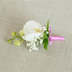 A refined floral boutonniere or corsage is shown resting horizontally on a light beige, linen-textured fabric that feels like a freshly pressed tablecloth laid out before a celebration in Mill Hill. The gently woven background, with its subtle threads and soft shadows, creates a calm, neutral canvas that allows the flowers to take centre stage. At the core of the design, a single white Phalaenopsis orchid opens gracefully, its broad, velvety petals shaped like gentle wings. The orchid's centre reveals a careful play of colour: soft yellow tones with just a whisper of pale pink around the throat, giving the bloom a warm, almost glowing heart. Nestled closely around the base of this central flower are airy clusters of white baby's breath, each tiny blossom forming part of a cloud-like surround that lends lightness, romance, and texture to the piece. A few slender, vivid green leaves slip out from beneath the blooms, offering a fresh, natural contrast to the white petals and the neutral backdrop. Off to one side, several closed, lime-green orchid buds sit along a slim stem, their compact forms hinting at blossoms that will unfurl later, suggesting continuity, growth, and the unfolding of a special day. The stems of the entire arrangement are tightly bound with a bright fuchsia satin ribbon. The ribbon is tied into a neat, small bow, its smooth, glossy surface catching the light and adding a joyful splash of colour that feels perfect for a wedding at Mill Hill Synagogue or a prom at a nearby school. The lighting is soft and even, revealing every delicate petal edge, tiny baby's breath flower, and the gentle sheen of the fabric and ribbon, resulting in an image that conveys quiet anticipation and elegant celebration. Designed with the care a local florist from Flowers Mill Hill would bring to each order, it looks ideal for placing on a lapel, wrist, or clutch bag for a formal event or intimate celebration.