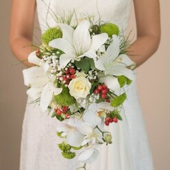 Cradled lovingly in the hands of a Mill Hill bride, this elaborate cascading bouquet tells a vibrant story of romance and celebration. Creamy white oriental lilies take centre stage, their petals arching outward and marked by faint cinnamon-speckled detail, while a tightly wound cream-hued rose sits at the heart, radiating classic beauty so often sought after in Mill Hill's wedding traditions. The arrangement bursts with contemporary fresh energy thanks to lime-green pom-pom chrysanthemums, their playful texture inviting the eye and conjuring memories of morning strolls in Mill Hill Park. Interwoven among these blooms are clusters of glistening red hypericum berries, offering pops of festive colour that echo the spirit of joyful gatherings on Mill Hill's high streets. Delicate sprays of white baby's breath fill in the spaces with airy lightness, further enhanced by wisps of fine greenery and the trailing elegance of white Phalaenopsis orchids, whose blooms seem to dance in the afternoon breeze. Nestled against the subtle lace of the bride's dress, the bouquet becomes a work of living art-each flower and berry meticulously selected for its vibrancy and meaning. As daylight floods into the scene, every nuance-from the satiny texture of petals to the dew-bright shimmer of the berries-is brought into sharp focus. This Mill Hill-crafted bouquet embodies both grace and joyful energy, capturing the essence of wedding floristry beloved by couples across North London.