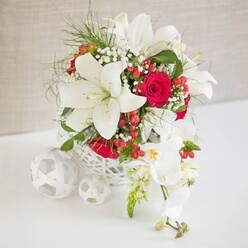 An elegant wedding-style table arrangement is captured in soft, even light, set against a pale beige textured backdrop that mirrors the calm, neutral tones of many Mill Hill homes. At the centre sits a low, round container crafted from white open-weave material, resembling a finely woven basket that lends the display a natural, artisanal character. From this airy base, large white oriental lilies rise and open, their sculptural petals spreading wide to reveal tall stamens tipped with warm pollen, giving the flowers a luminous, almost star-like quality. Intertwined with the lilies are rich red roses, velvety and full, each bloom tightly layered and deep in colour, adding a romantic intensity that would feel at home in intimate gatherings near Mill Hill Broadway. Throughout the arrangement, delicate sprigs of white baby's breath thread their way between the larger flowers, creating a soft, misty texture and gentle transitions between tones. Slender, bright green foliage, reminiscent of fine ferns, loops and curves around the blooms, bringing freshness and subtle movement as if the bouquet were freshly arranged for delivery across Mill Hill and neighbouring suburbs. Small clusters of glossy red hypericum berries punctuate the design with jewel-like accents, enhancing the festive interplay between red and white. On the right, a graceful cascade of white phalaenopsis orchids pours over the basket's edge, each orchid bloom bearing smooth petals and a hint of yellow and pink in the centre, while a few buds at the tip add a sense of unfolding time. To the left, three decorative white woven spheres rest on the white tabletop, mirroring the container's texture and completing the balanced, modern composition. The overall impression is of refined celebration and gentle opulence, perfectly suited to wedding receptions, engagement dinners, or seasonal gatherings in Mill Hill.