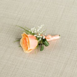 Placed delicately across a pale beige, linen-textured fabric, this peach rose boutonniere looks as though it has just been set down on a dressing table before a Mill Hill ceremony. The background fabric has a fine, even weave, soft and slightly matte, which gently catches the light and allows the flowers to stand out without distraction. At the heart of the arrangement is a single rose in a soft peach tone, its petals opening in a graceful spiral from a deeper, warm apricot centre toward lighter, almost cream-tinted outer edges. The rose appears freshly cut, with petals that look velvety to the touch and give off the impression of a subtle, sweet scent. Clustered near its upper right are tiny heads of white baby's breath, their airy, cloud-like sprays bringing a sense of romance reminiscent of classic North West London weddings. Slender green stems and fine, grass-like fronds of foliage arc gently around the main bloom, while two broader, dark green leaves curl neatly near the base, adding structure and a deeper tonal anchor. The stems are expertly wrapped in a satin peach ribbon, tightly coiled and finished with a small, precise bow that mirrors the rose's soft hue. The overall lighting is even and warm, highlighting the textures of the petals, foliage, and ribbon while casting only the faintest shadows. This buttonhole feels perfectly suited for a groom or groomsman heading to a celebration near Mill Hill Park or arriving for photos on a quiet residential street, offering a look of understated sophistication and the tender charm of new beginnings.