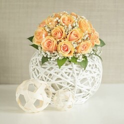 An elegant floral display features a generous cluster of around two dozen peach and apricot roses, carefully arranged into a smooth, domed shape that immediately draws the eye, like a glowing focal point in a Mill Hill living room flooded with afternoon light. Each rose is tightly layered, with deeper, almost burnt-orange centers that gradually fade to softer, buttery edges, giving the bouquet a warm, romantic radiance. Between the roses, tiny white blooms of baby's breath thread in and out, creating a misty, cloud-like veil that lightens the palette and adds a touch of relaxed sophistication, perfect for a wedding table near Mill Hill East station or an anniversary dinner set up in a quiet NW7 townhouse. Dark green leaves, mostly hidden but occasionally visible near the base, anchor the softness of the flowers and offer a subtle, natural contrast. The arrangement is cradled in a round white vessel formed from intricately woven strands, resembling a delicate wicker orb; its open lattice structure lets you see just enough of the interior to appreciate the craftsmanship without distracting from the flowers. In front of this main orb, two smaller decorative balls echo the same woven style: one larger and patterned like a minimalist soccer ball, the other more loosely formed, adding sculptural interest to the clean white tabletop. Behind it all, a pale, beige-grey wall with the gentlest texture creates a calm, uncluttered backdrop that enhances the sense of serenity. The soft, even lighting suggests a carefully styled interior scene, the sort of centerpiece a local florist from Flowers Mill Hill might design for same-day flower delivery to homes and venues across the Mill Hill Broadway and Daws Lane area.