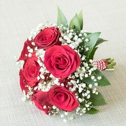 Bathed in soft daylight filtered through a nearby window, a compact bouquet of sumptuous red roses and airy baby's breath rests on an off-white, linen-textured surface, reminiscent of the calm, inviting interiors found in Mill Hill's local cafés. Each rose, its petals arranged in a perfect spiral and glowing with deep, velvety crimson, speaks of enduring love and classic romance. Interspersed among the roses, delicate sprays of white gypsophila lend a cloud-like lightness and purity to the design, creating a gentle interplay of colour and texture. Dark, emerald-green leaves, likely ruscus or salal, add a lush framing touch, their glossy surfaces reminiscent of the well-tended greenery along Daws Lane. At the bouquet's base, the sturdy stems are carefully wrapped in a striking red and white striped ribbon, elegantly spiraled with glistening white pearls-details that hint at the expert hands of Mill Hill's finest florists. The composition radiates warmth, making it an iconic choice for weddings, romantic gestures, or heartfelt thank-you bouquets, each petal and bead exuding a sense of celebration and community unique to Mill Hill.