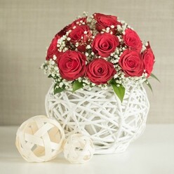 This image presents an intimate wedding table arrangement that could easily grace a reception venue or family home in Mill Hill. The central focus is a lush, dome-shaped cluster of deep crimson roses, perhaps fifteen blooms gathered so tightly that their spiralled petals touch, creating a plush, cushion-like surface of colour. Each rose is open at just the right stage, full yet not overblown, suggesting careful selection by an experienced florist. Intermingled with the roses are light sprays of white gypsophila, the tiny blossoms appearing like a dusting of soft snow across the bouquet. Their brightness lifts the darker reds and introduces a feeling of air and space, while a few discreet green leaves peek from within, bringing a subtle hint of foliage and natural structure. The flowers are cradled in a distinctive round vase formed from white, interwoven strands that twist and cross to create an open, nest-like design, its bulbous shape echoing the rounded curve of the rose dome. The white of the vase reflects gentle, even light, reinforcing the freshness and clarity of the composition, much like the clean lines of modern homes along Lawrence Street or on the leafy slopes near Mill Hill Broadway. To the left of the vase, two decorative spheres made from light, woven material in cream and beige tones rest on the same smooth, pale surface, their airy construction mirroring the latticework of the vase and adding a touch of sculptural interest. The background is a softly blurred, neutral wall that recedes quietly, ensuring that all attention rests on the roses and textured whites. The arrangement evokes warmth, romance, and thoughtful hospitality, suitable for an intimate dinner, wedding breakfast, or same-day flower delivery to a Mill Hill address.