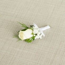 A classic white rose boutonniere is displayed on a diagonal across a neutral, light beige background that looks like finely woven linen, the kind of fabric you might find laid out on an elegant Mill Hill reception table. The central rosebud is a creamy, soft ivory, its petals tightly layered and velvety, holding the promise of full bloom as if timed perfectly for the moment a groom steps into his ceremony. Surrounding the rose are delicate clouds of white baby's breath, each tiny flower forming a star-like point of light that adds airiness and romance to the arrangement. The greenery is a mix of slender, dark green leaves and a few feathery, fern-like fronds, carefully placed to cradle the rose and give the boutonniere a natural, slightly woodland charm reminiscent of green corners around Mill Hill Park. All the stems are gathered and wrapped in smooth white satin ribbon, the binding impeccably straight and finished with a petite bow that sits just below the flower heads, signalling that this piece is ready to be pinned to a suit or tuxedo lapel. The light in the image is soft and diffused, casting no harsh shadows, but instead tracing the gentle curves of each petal, the fine texture of the gypsophila, and the subtle sheen of the ribbon. This boutonniere radiates understated sophistication and is perfectly suited for weddings, civil ceremonies, or formal events across Mill Hill, from intimate gatherings at local venues to more elaborate celebrations, capturing the tenderness and excitement of a day built around love and shared vows.