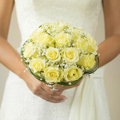 Under the gentle embrace of soft lighting, this Mill Hill bridal bouquet radiates serene beauty and expert floristry. The centerpiece consists of tightly clustered, pale yellow roses, their velvety petals curling outward in a harmonious swirl, creating a plush, domed silhouette. Accentuating the arrangement, airy sprays of baby's breath weave through the roses, adding delicate contrast and whisper-light texture-recalling the tranquil fields and blooming gardens of Mill Hill village. Graceful ribbons of deep green foliage, perhaps artfully shaped wire, frame the base, lending structure reminiscent of Mill Hill's well-kept hedges and local parks. The bride's hands are visible, nails polished a subtle pink, gently grasping the stems and showcasing a glimmer of a wedding ring. Her white gown, richly textured, acts as an elegant canvas for the bouquet's cheerful hues. Subdued, blurred tones in the background ensure the florals remain the captivating focal point, creating an atmosphere both intimate and celebratory. This image reflects the delightful balance of tradition and modern beauty found in Mill Hill weddings, offering inspiration for those envisioning a bouquet that embodies freshness, hope, and a touch of local charm.