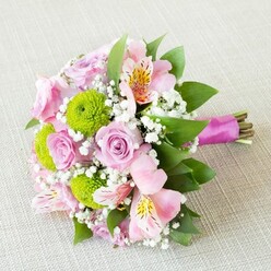 This hand-crafted bouquet, a celebration of fresh beginnings, rests serenely on a light beige woven surface-a fitting tableau for moments cherished in Mill Hill. The arrangement showcases velvet-soft pink roses, their spiral centers and gradations of bloom stages echoing the elegant gardens framing St. Joseph's College. Scattered amid the pastel roses are vivid lime button chrysanthemums, whose firm, spherical blossoms enliven the bouquet with modern vitality. Pink alstroemeria unfurl in blushing tones, each petal lightly dusted with gold and maroon markings resembling a subtle painter's flourish. Sprays of white gypsophila evoke delicate wisps of morning mist settling over Mill Hill's country walks. Deep, lush green foliage weaves throughout, providing structure and a whisper of the area's verdant surroundings. A rich magenta ribbon secures the stems, adding a vibrant finishing note and quietly reflecting the creative, neighbourly spirit of local events near Mill Hill Broadway. Every floral element is gently illuminated by balanced light, revealing crisp edges and the tactile joys of petal and fabric. This exquisite bouquet, ideal for bridesmaids, romantic gifts, or special ceremonies in NW7, offers a fresh breath of floral beauty-a heartfelt gesture crafted for life's most memorable occasions across Mill Hill.