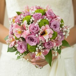 Nestled in the gentle hands of a bride against the ornate lace of her wedding dress, this Mill Hill bridal bouquet captures an exquisite blend of tradition and modern flair. The floral arrangement exudes romance, with graceful roses in soft pink and pale lavender, their velvet petals unfolding to reveal subtle hints of fragrance reminiscent of spring mornings in the NW7 gardens. Amongst the roses, vibrant lime green button chrysanthemums offer a lively contrast, their compact, globe-shaped blossoms adding a contemporary spark to the palette. Pink alstroemeria lilies introduce intricate maroon and yellow markings, peeking out with an exotic elegance that suggests wild beauty. Throughout the bouquet, baby's breath trails in delicate, white clouds, softening and elevating the whole composition with breezy lightness. Deep green leaves, possibly from the roses themselves, frame and support the bouquet, grounding its pastel blooms with rich, natural depth. Every element is carefully placed yet relaxed, echoing Mill Hill's balance of heritage and innovation. The overall impression is one of fresh beginnings, designed by a local florist passionate about bringing the joy of flowers to weddings and special celebrations across Mill Hill and its welcoming community.