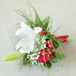Resting atop a finely woven, pale beige fabric reminiscent of Mill Hill's timeless interiors, this floral arrangement is a testament to local craftsmanship and thoughtful design. The bouquet's star, a fully open white lily, commands attention with its broad, snowy petals delicately dotted with subtle copper specks and prominent yellow anthers, casting an aura of freshness and grace. Tucked in close, a rich, red rose radiates intensity and warmth, its perfectly curled petals conjuring the romance of a summer evening in Mill Hill Park. Wisps of airy baby's breath impart an ethereal touch, their tiny, starry blossoms lifting the visual weight and brightening the palette. Clusters of shiny, crimson berries nestle between the blooms, breathing a festive accent into the composition, reminiscent of neighbourhood celebrations. On the left, a slender green lily bud stretches upward, symbolising the promise of blossoming friendships and new beginnings in North London. The ensemble is framed by an artful mix of feathery fern and lush, deeper green leaves, evoking natural woodland borders. Finished with a luxurious red satin ribbon, the bouquet's stems are gathered with care, the ribbon glinting in the light and tying together the palette beautifully. This bouquet feels celebratory yet understated-ideal for weddings, anniversaries, or heartfelt gestures, hand-delivered by trusted Mill Hill florists.