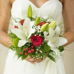 Bathed in the soft afternoon light of Mill Hill, this close view finds a bride in a white, subtly patterned gown, holding a bouquet brimming with emotional and botanical depth. Central to the display are luminous oriental lilies, their snowy petals open and radiant, exuding purity and hope for the journey ahead. Tucked among them, deep red roses coil tightly-their rich color infusing the arrangement with an undercurrent of timeless romance. Ruffled sprays of white baby's breath weave delicately through the bouquet, while bright green lily buds hint at future joys yet to blossom. Sprightly clusters of red hypericum berries and mossy green button blooms lend playful pops of color and natural movement, reminiscent of explorations through Mill Hill's beautiful parks. Framing it all is a lush medley of greenery-from airy ferns to broad, classic leaves-anchoring the bouquet with texture and natural elegance. The bride's hands, gently clasping the stems, add to the gentle sense of anticipation. Created by a knowledgeable Mill Hill florist, this vibrant arrangement offers both a tactile and visual feast for the senses-its colours, textures, and design capturing the heartfelt spirit of a North London wedding. More than flowers, it embodies a story of love, commitment, and memory, destined to become a highlight of the day's celebrations.