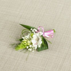 A refined floral boutonniere lies against a pale beige, linen-textured fabric, the understated background echoing the calm, residential charm of Mill Hill's quiet streets. At its centre, a white alstroemeria opens gracefully, the petals lightly ruffled at the edges and delicately patterned with fine reddish-brown flecks that draw the eye inward to the soft, pollen-dusted stamens. Just to one side, a compact, cone-shaped spike of vivid green buds, reminiscent of Ornithogalum, provides an intriguing contrast, its tightly layered structure adding a contemporary touch to this otherwise classic piece. Threaded between these elements are airy sprigs of white baby's breath, each tiny flower like a pinprick of light, creating a cloud-like halo that softens the overall look and makes the design feel weightless. The base is formed from sleek, dark green, sword-like leaves that ground the composition, while wisps of fine, grass-like foliage spill out in gentle curves, suggesting movement and a sense of organic growth. The stems are bound neatly at the right-hand side with a bright pink satin ribbon, tied in a precise, small bow whose sheen catches the light, adding a sweet, celebratory accent to the otherwise calm palette of whites and greens. The boutonniere is photographed in soft, even lighting that reveals every texture, from the smoothness of the petals to the crisp line of each leaf. This intimate close-up captures the quiet elegance of a piece that could be worn to a Mill Hill wedding at a local church or registry office, a prom celebration, or an anniversary dinner in one of the area's cosy restaurants, radiating fresh, understated romance.