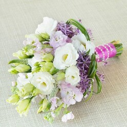 Delicately arranged with heartfelt artistry, this bouquet appears poised atop a neutral, cream-beige fabric-reminiscent of the understated luxury and warmth found within Mill Hill's finest local venues. Flourishing white lisianthus blooms, round and full, open delicately against gathering buds of chartreuse and lime, symbolising fresh hope and quiet anticipation. Wispy clusters of lavender-pink stock flowers weave gentle threads of perfume through the arrangement, their airy blooms bringing a touch of meadow softness like a springtime stroll through Mill Hill Park. Striking deep purple allium heads, bursting with star-shaped florets, create bold accents-mirroring bursts of colour from local gardens at the height of summer. Sleek bear grass blades arc gracefully overhead, adding motion and verdant zest, while light green buds and textured fillers create subtle yet lively depth. The base is finished in bright fuchsia satin ribbon that draws the eye, topped with a delicate overlay of gleaming white pearls-a modern twist on classic bridal charm. This radiant bouquet, reflecting both romance and celebration, is destined for joyful gatherings by the sacred green spaces and community halls of Mill Hill, and makes the perfect muse for stories of local love and floral artistry.
