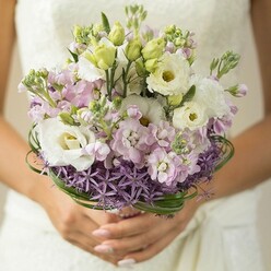 In the soft, crisp air of Mill Hill, this wedding bouquet becomes a true expression of joy and artistry, lovingly cupped in a bride's hands. White Lisianthus flowers, their petals unfurling in delicate ruffles, radiate gentle light, flanked by unopened green buds that add a whisper of anticipation. Clusters of light pink and lilac stock create layers of texture and gentle movement, like the shifting blooms along Arrandene Open Space. At the orbit of the bouquet, a dramatic halo of deep purple, star-shaped Allium provides a captivating contrast, grounding the pastel blooms above with bold, sculptural flair similar to the ornamental displays along Mill Hill Broadway. Loops of slender bear grass arc gracefully around the bouquet's midsection, providing green structure while echoing the area's winding leafy lanes. The bride's hands are polished in a subtle nude shade, their gentle hold reinforcing the careful craftsmanship within each flower's placement. Behind the bouquet, a blurred hint of lace or textured ivory from the bride's dress completes the image, drawing out an air of pure celebration. This bouquet, fresh from Flowers Mill Hill and available throughout NW7, brings together modern romance and timeless elegance-an emblem of love flourishing in a classic London neighbourhood.