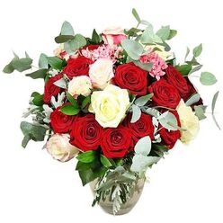 Displayed against a pure white background, this luxurious rose bouquet in a clear glass vase immediately draws the eye with its intense colour and carefully balanced form. A profusion of deep red and vivid scarlet roses, easily two dozen or more, form the heart of the arrangement, their velvety petals spiralling outward in soft layers that suggest deep affection and classic romance. Some roses remain in tight bud, hinting at emotions still unfolding, while others are fully open, revealing generous, plush centres. Nestled between these passionate tones, creamy white and pale ivory roses, along with a few white blooms edged with faint blush-pink, lend a gentle glow and serenity, creating visual breathing space and an air of refinement. Threaded through the larger flowers, tiny clusters of bright pink filler blossoms add lively texture and a brighter, playful note without disturbing the overall elegance. Encircling and weaving among the blooms, blue-green eucalyptus branches with their rounded, slightly silvery leaves provide a fresh, modern counterpoint and evoke the idea of a light, soothing fragrance. Deeper green foliage and a scattering of silvery-green leaves support the composition, shaping it into a full, rounded profile that appears lush from every angle. The vase, with its softly rounded base and slimmer neck, reveals straight stems and clear water, giving a sense of honesty and freshness to the abundant display. This arrangement would look perfectly at home in a Mill Hill living room overlooking the green spaces near Lawrence Street, or as a same-day delivery from Flowers Mill Hill to a couple celebrating an engagement close to Arrandene Open Space, symbolising love, joy, and shared new beginnings.