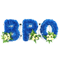 Presented on a crisp white backdrop, this three-letter floral tribute spells out BRO in substantial, upright forms crafted entirely from royal blue chrysanthemums. The surface of each letter appears tactile and plush, with thousands of small blue florets pressed tightly together to create seamless curves and sharp, defined edges. Light caresses the blooms, highlighting subtle variations in petal sheen that make the blue feel simultaneously rich and tranquil. Each letter carries a small, carefully placed spray of cream to pale yellow roses and tiny white fillers - possibly mini chrysanthemums or baby's breath - arranged near the base: the B's cluster at the lower left and matching accents at the lower right of the R and O. Interspersed among the flowers are deeper green, glossy leaves and lighter, fern-like foliage that give the design structure and a gentle, aromatic freshness that lingers when you approach. A slim white foam or base is visible at the letter perimeters and in the openings of the B, R and O, revealing the precise florist construction behind the sentimental surface. The piece maintains a solemn, respectful mood, ideal as a personalised funeral tribute for a brother or close companion: the blue suggests steadfast remembrance, the pale roses a soft consolation. Imagine this arranged by a local Mill Hill florist and placed near a service at a neighbourhood chapel or brought to a family home on The Ridgeway as a quiet, dignified sign of sympathy.