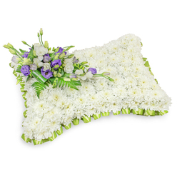 A reverent floral pillow arranged on a pristine white background, formed almost entirely from hundreds of small, immaculate white chrysanthemum heads that create a seamless, cushion-like surface. The dense chrysanthemums give the piece a soft, cloudlike texture - a symbol of peace and repose - while a subtle drop shadow beneath confirms its gentle three-dimensional presence. Encircling the rectangular form, a pale green ribbon has been meticulously pleated into an even, scalloped border, the fresh lime tone offering a quiet contrast that suggests renewal amid mourning. Diagonally across the top-left sits a carefully gathered spray of blooms: layered white lisianthus share space with deep purple lisianthus, some in bud and some opened to reveal their ruffled throats, providing a respectful color accent that signals dignity and remembrance. Sprays of delicate fern fronds and glossy green foliage are woven through this cluster, adding texture and a living brightness, while a dark green leaf wrap secures the base with clean, understated craftsmanship. The overall impression, captured under soft studio lighting, is one of serene comfort-an arrangement well suited to funeral services, graveside tributes, or moments of private condolence across Mill Hill. As a local florist we present this tender composition with the same careful attention given to every memorial delivery across Mill Hill Broadway and surrounding streets, offering a quiet language of flowers when words fall short.
