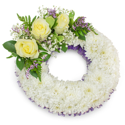 An elegant circular funeral wreath assembled from a plush mass of immaculate white chrysanthemums, each bloom contributing a feathery, layered texture that forms a sympathetic, cloud-like ring. The abundant chrysanthemum heads are composed so tightly that the surface appears almost like white linen-soft to the eye and resolutely dignified. Subtle threads of deep lavender-purple mesh or ribbon peek through the flowers along both the inner and outer edges, introducing a refined line of colour that gives the piece quiet depth. Positioned on the upper-left quadrant is a gentle focal spray: three soft, buttery-yellow roses, their petals opening with a restrained grace, convey affection and remembrance. These roses are cushioned by sprays of tiny white gypsophila-baby's breath-whose tiny sparkles of white lightness temper the solidity of the chrysanthemums, while small clusters of pale purple statice lend a whisper of seasonal violet. A harmonious mixture of lush green foliage-some broad and glossy, some finely textured like fern fronds-frames the accent cluster and lifts the composition's overall freshness. The wreath maintains a perfect circular shape with a clear centre, symbolic of unbroken bonds and continuity. Photographed on a stark white surface with soft, diffuse lighting, the bouquet casts a faint shadow that gives it dimensional presence. As if prepared by a florist who knows Mill Hill's quieter streets and memorial traditions, this wreath offers a tasteful, comforting expression of condolence suited to funerals, wakes, and graveside services across NW7 and nearby Mill Hill East.