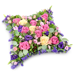 This image presents an elegant funeral cushion, styled as a floral pillow that appears both soft and reassuring, as though placed gently beside a coffin or memorial stone in Mill Hill. The arrangement forms a low, rectangular cushion with subtly rounded corners, the entire surface closely planted with flowers so that no spaces are left bare. Large roses in tones of vivid fuchsia, gentle pastel pink, and soothing cream or pale yellow dominate the design, many of them fully open, their petal layers overlapping in delicate spirals that give a sense of luxury and tenderness. Woven among these central blooms are rich purple lisianthus, their deep colour introducing a thoughtful, contemplative mood and a velvety, ruffled texture. Between the larger flowers, clusters of tiny lavender-toned filler blossoms, reminiscent of statice or limonium, add intricacy and a light, airy feel, while petite pink spray roses provide additional softness and intimacy. Supporting the flowers is a generous layer of foliage: rounded, silvery-green eucalyptus leaves lend a cool, calming presence, while darker, fern-like greenery brings depth and a more natural, garden-inspired character, as if gathered from the green spaces around Mill Hill Park. The arrangement is displayed against a plain white background, a subtle shadow beneath it suggesting its thickness and cushion-like form. The lighting is gentle and even, allowing the pinks, creams, and purples to glow without harsh contrast. Overall, the floral pillow expresses sympathy and solace, a tender tribute that speaks quietly of love, remembrance, and the wish for peaceful rest, suitable for funeral services and commemorative gatherings throughout the Mill Hill community.