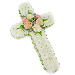 A traditional Christian cross fashioned entirely from fresh flowers is presented against a bright white background, its dimensional edges defined by a faint yet distinct shadow. The structure of the cross is hidden beneath an immaculate covering of white button chrysanthemums, each tiny blossom carefully placed so that the entire surface appears smooth and consistent. This dense blanket of crisp white blooms suggests purity, peace, and a gentle, prayerful atmosphere, much like the hush that settles over Mill Hill's churches before a service begins. Just at the edge of the cross, a slim line of lime green ribbon traces the outline. The ribbon's fresh, almost citrus shade peeks out beneath the rim of petals, giving the piece a refined, finished appearance and adding a subtle splash of colour that hints at renewal and hope. At the intersection of the cross's arms lies the most intricate detail: a compact gathering of roses and foliage. Here, fully open roses in creamy white, soft blush pink, and warm peach tones nestle together, their velvety petals unfolding in gentle spirals. Surrounding and threading between them are arching fronds of rich green fern and dainty spikes of lavender-coloured filler flowers, which introduce a soft purple accent to the otherwise white and green palette. This central cluster acts as the emotional heart of the tribute, drawing the eye and suggesting love, remembrance, and gratitude. Designed as a sympathy arrangement suitable for delivery to local Mill Hill chapels, crematoriums, or family homes, the floral cross communicates respect, faith, and a tender farewell in a quietly elegant and deeply comforting way.
